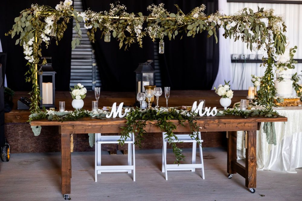 Una mesa de novios rústica bajo un dosel de eucalipto y flores blancas, sobre madera al natural