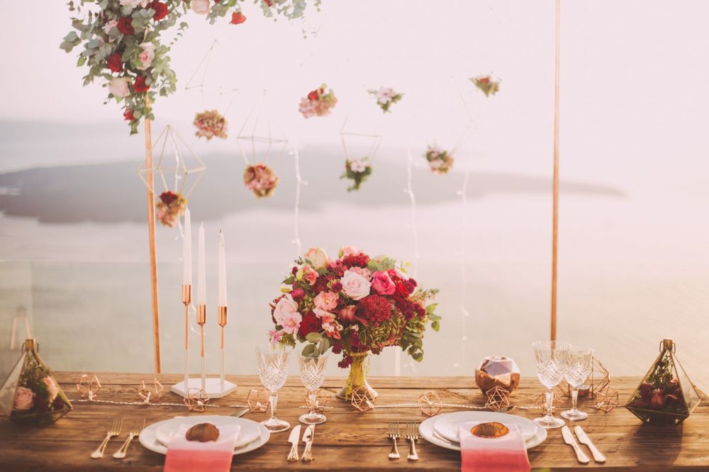 Una mesa al borde de un acantilado al atardecer con un ramo de rosas en tonos baya intensos y rosa empolvado, candelabros de cobre y vista al mar