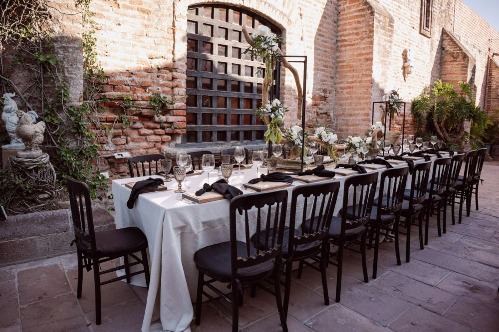 Una mesa al aire libre frente a un cálido muro de ladrillo terracota, con lino crema y sillas negras