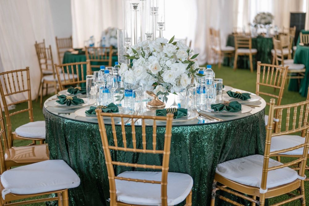 Una mesa de recepción redonda con mantel esmeralda intenso, sillas doradas y un centro generoso de rosas blancas